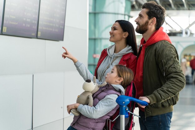 Zijaanzicht van de gelukkige ouders en hun dochter die voor het tijdschemabord staan terwijl ze wachten op de vlucht op de luchthaven. reizend concept. stock foto