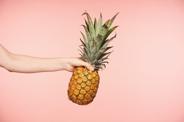 Zijaanzicht van de hand van de mooie vrouw met naakte manicure die vingers samenknijpt terwijl grote verse ananas wordt vastgehouden, die tegen roze achtergrond wordt geïsoleerd