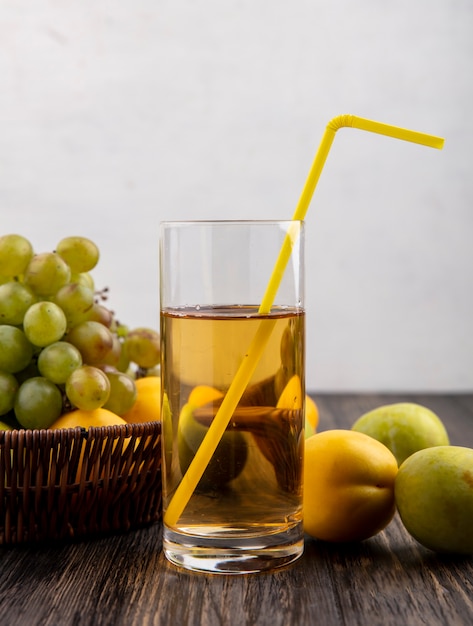 Zijaanzicht van druivensap met drinkbuis in glas en mandje van druivennectacot met plukken op houten oppervlak en witte achtergrond