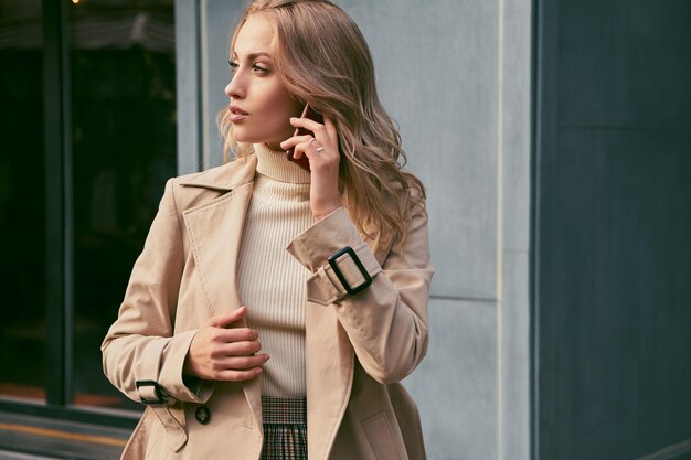 Zijaanzicht van een aantrekkelijk, casual blond meisje in een trenchcoat die op de mobiel praat en dromerig naar buiten kijkt