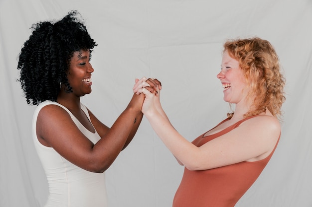 Zijaanzicht van een Afrikaanse en blonde jonge vrouw die elkaars hand houden tegen grijze achtergrond