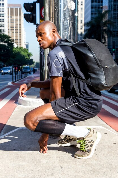 Zijaanzicht van een Afrikaanse jonge sportieve man met zijn rugzak gehurkt in het midden van de weg