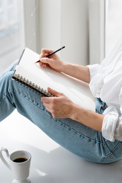 Zijaanzicht van een jonge vrouw die thuis dichtbij het raam tekent