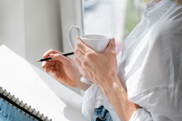 Zijaanzicht van een jonge vrouw die thuis dichtbij het raam tekent