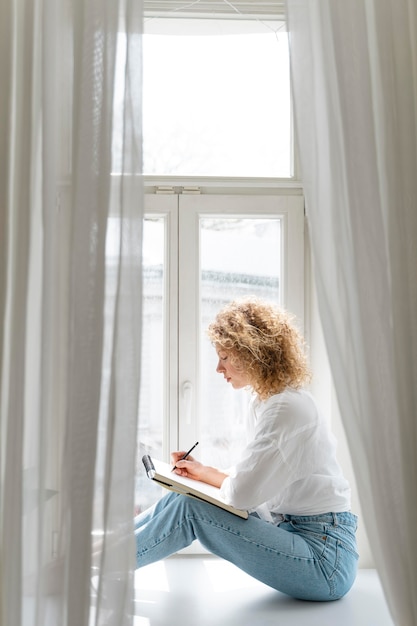Gratis foto zijaanzicht van een jonge vrouw die thuis dichtbij het raam tekent