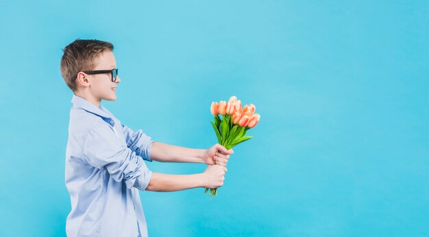 Zijaanzicht van een jongen die oogglazen draagt die verse tulpen geven tegen blauwe achtergrond