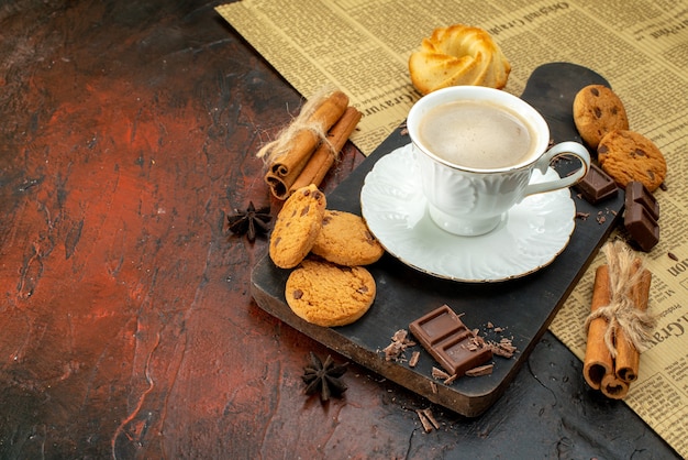 Zijaanzicht van een kopje koffie op een houten snijplank op een oude krant, koekjes, kaneellimoenen, chocoladerepen op donkere achtergrond