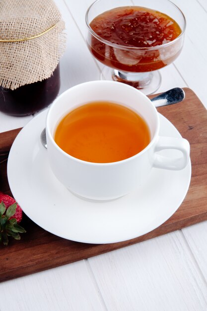 Zijaanzicht van een kopje thee met aardbeienjam in een glazen vaas op houten bord op wit