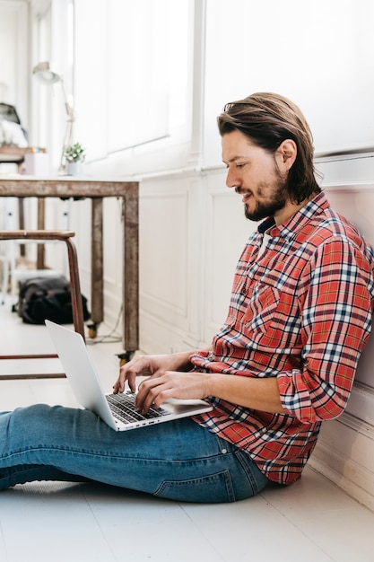 Zijaanzicht van een lachende jonge man zittend op hardhouten vloer te typen op laptop