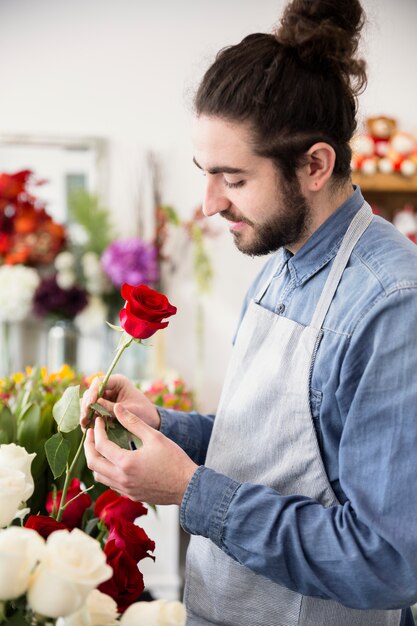 Zijaanzicht van een mannelijke bloemist die rode roze bloem in zijn bloemwinkel bekijkt