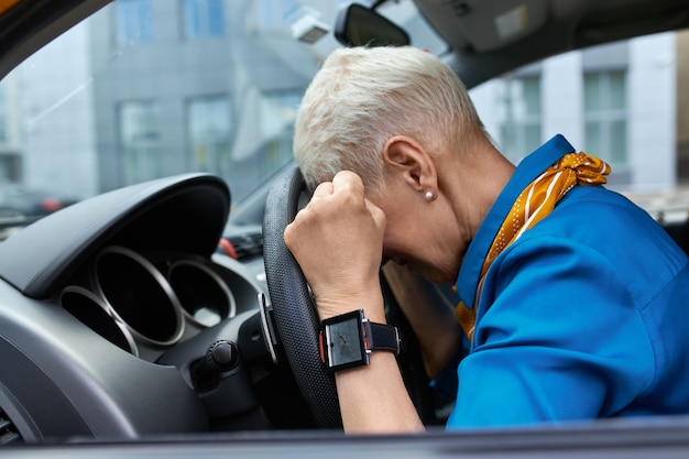 Zijaanzicht van een ongelukkige gestreste vrouw van middelbare leeftijd knijpen vuisten en hoofd rustend op het stuur, vast in de file, te laat komen op het werk of een auto-ongeluk krijgen, zittend op de bestuurdersstoel