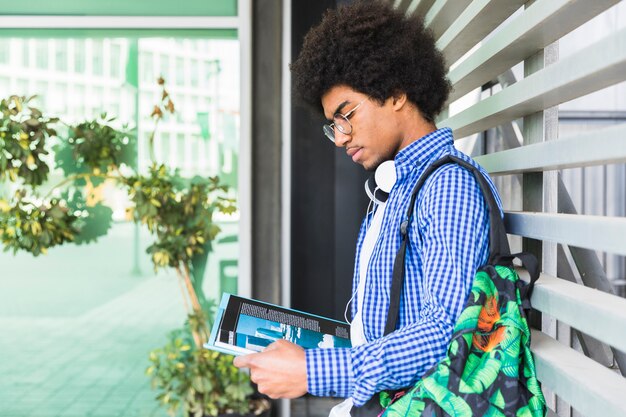 Zijaanzicht van een tiener mannelijke student draagtas op zijn schouder leunend tegen muur lezen van het boek