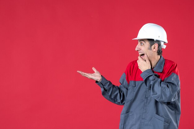 Zijaanzicht van een verraste jonge werknemer in uniform met een helm die iets aan de rechterkant wijst op een geïsoleerde rode muur