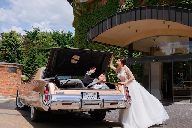 Gratis foto zijaanzicht van een vrolijke bruid in een mooie trouwjurk die tegenover de retro-auto op het hotelgebied staat en zich leuk voelt tijdens het sluiten