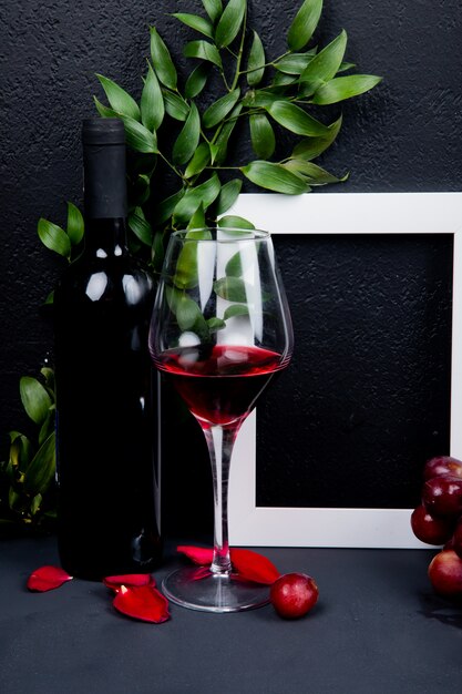 Zijaanzicht van fles en glas rode wijn met frame en bladeren met bloemblaadjes op zwart met exemplaarruimte