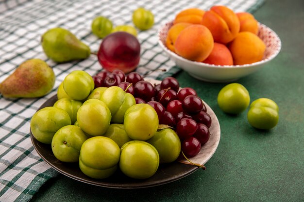 Zijaanzicht van fruit als pruimen, kersen, abrikozen in plaat en kom met peer en perzik op geruite doek op groene achtergrond