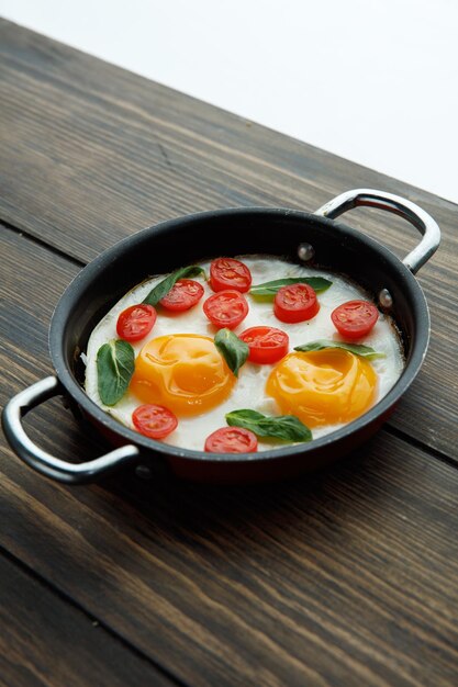 Zijaanzicht van gebakken eieren met tomaten in koekenpan versierd met bladeren op houten achtergrond
