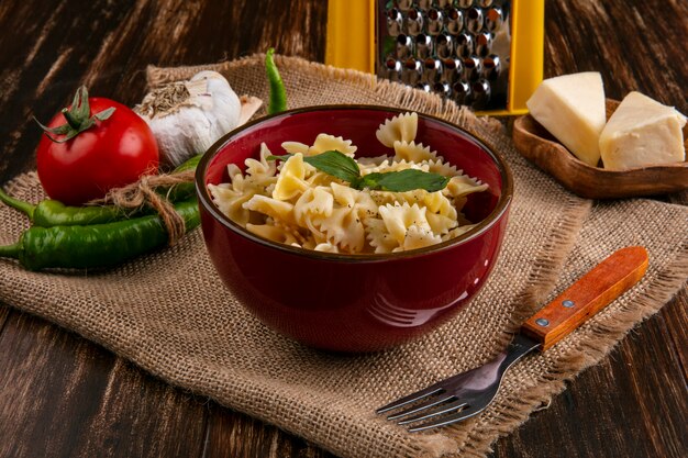 Zijaanzicht van gekookte pasta in een kom met een vork tomaten chili pepers knoflook en kaas op een beige servet