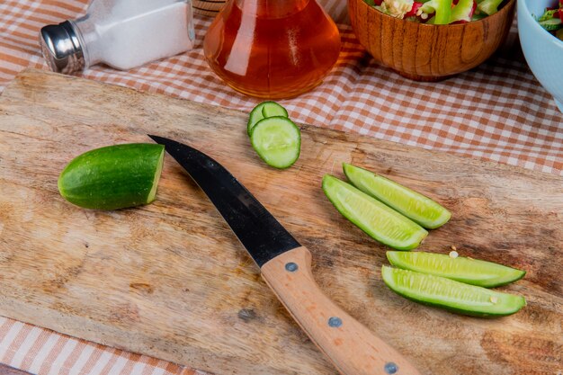 Zijaanzicht van gesneden en gesneden komkommer met mes op snijplank met plantaardige salade gesmolten boterzout op geruite doek