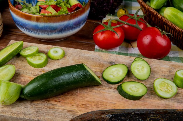 Zijaanzicht van gesneden en gesneden komkommer op snijplank met tomaten groente salade op houten tafel