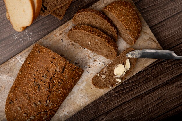 Zijaanzicht van gesneden en gesneden sandwichbrood en boter die op broodplak wordt uitgespreid met mes op scherpe raad op houten achtergrond