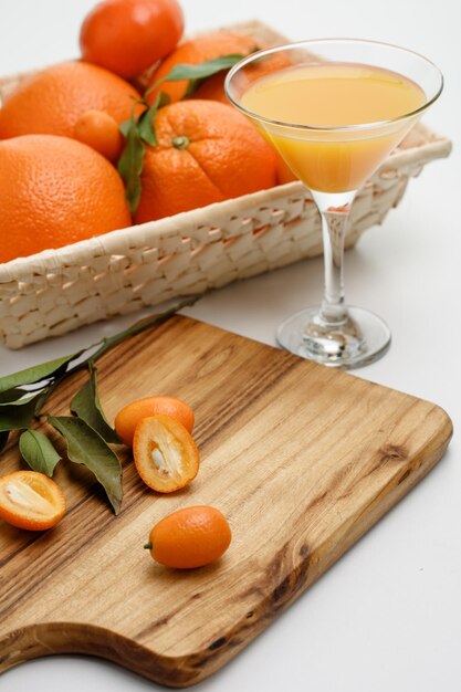 Zijaanzicht van gesneden en hele kumquats met bladeren op snijplank met mand met sinaasappels en kumquats en glas sinaasappelsap op witte achtergrond