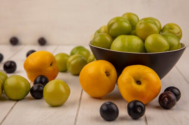 Zijaanzicht van groene kersenpruimen op een kom met fruit zoals perzikessloes die op een houten achtergrond worden geïsoleerd