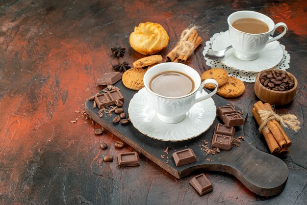 Zijaanzicht van heerlijke koffie in witte kopjes op houten snijplank koekjes kaneel limoenen chocoladerepen aan de linkerkant op gemengde kleur achtergrond