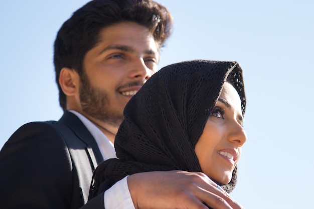 Zijaanzicht van jong romantisch koppel. Aantrekkelijke moslimvrouw in zwarte hoofddoek en knappe donkerharige man die haar omhelst terwijl ze dichtbij staat en ver weg kijkt. Romantiek en relatieconcept