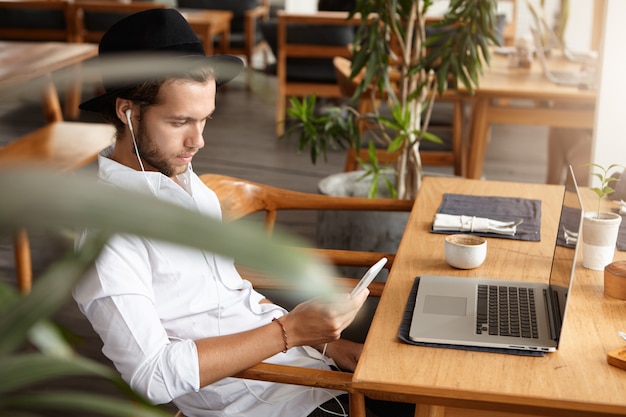 Gratis foto zijaanzicht van knappe jonge kaukasische freelancer of student zittend aan café tafel met open laptop pc, mobiele telefoon vasthouden en luisteren naar muziek op oortelefoons, met behulp van online app tijdens het ontbijt