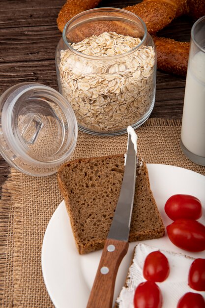 Zijaanzicht van kruik met haver-vlokken en plaat met roggebrood plak tomaten mes op zak op houten achtergrond