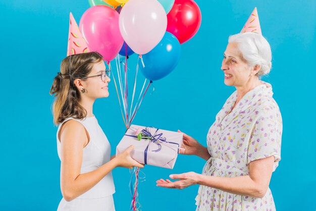 Zijaanzicht van meisje die verjaardagsgift geven aan haar gelukkige grootmoeder op blauwe achtergrond