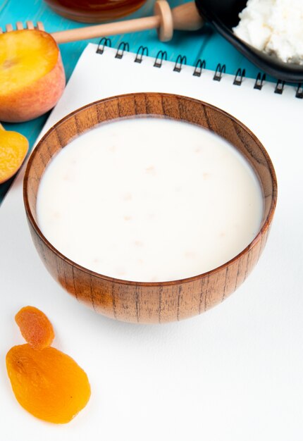 Zijaanzicht van melk in een houten kom op een schetsboek en gedroogde abrikozen verse rijpe perziken op blauw
