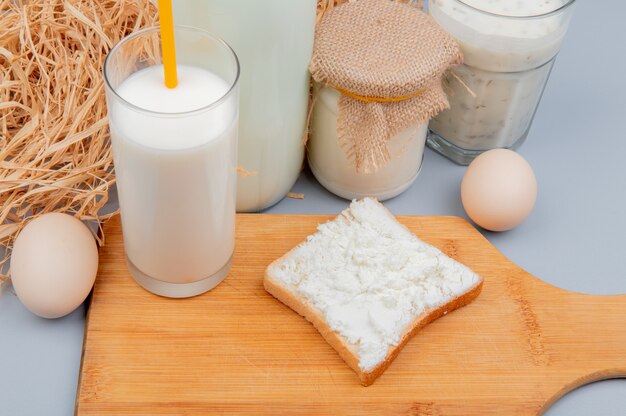 Zijaanzicht van melkproducten als kwark besmeurd met sneetje brood glas melk op snijplank room melk yoghurt soep en eieren met stro op blauwe oppervlak