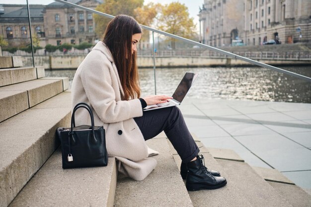 Zijaanzicht van mooi casual meisje dat op laptop werkt op trappen in de buurt van stadsrivier