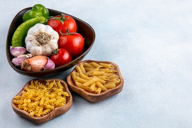 Zijaanzicht van ongekookte pasta in kommen met tomaten, knoflook en paprika in een kom op een grijze ondergrond