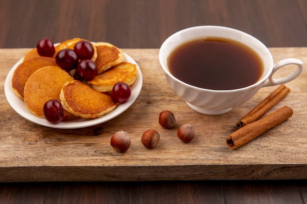 Zijaanzicht van pannenkoeken met kersen in plaat en kopje thee met kaneel en noten op snijplank op houten achtergrond