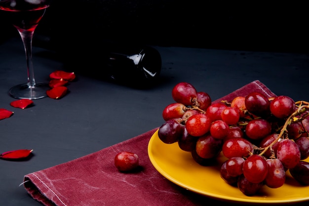 Zijaanzicht van plaat met druif op doek met glas en fles rode wijn met bloemblaadjes op zwarte