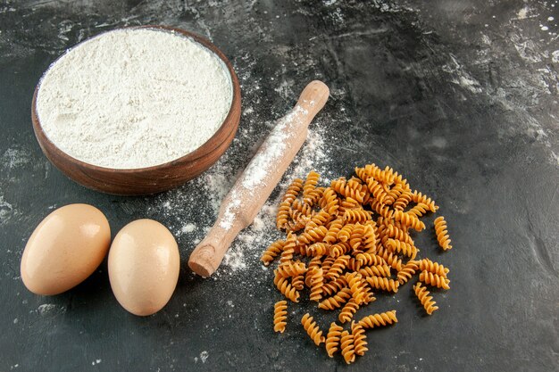 Zijaanzicht van rauwe Italiaanse pasta's twee eieren en bloem in een bruine kom deegroller op grijze achtergrond