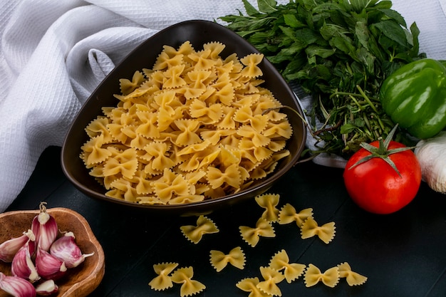 Zijaanzicht van rauwe pasta in een kom met tomaten, knoflook en paprika met munt op een witte handdoek op een zwarte ondergrond