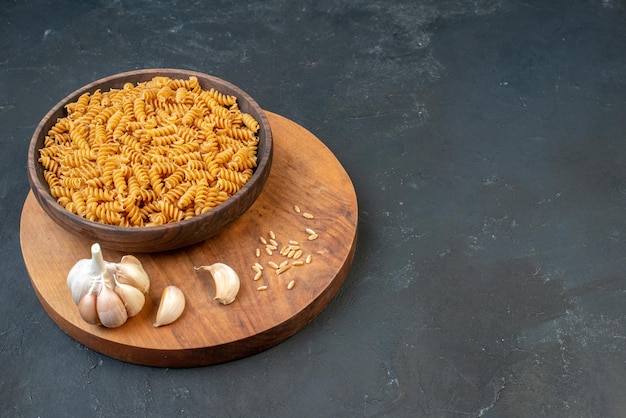 Zijaanzicht van rauwe pasta's in een bruine kom knoflookrijst op een houten bord aan de rechterkant op zwarte achtergrond