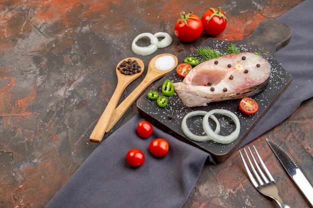 Zijaanzicht van rauwe vissen en peper-ui greens tomaten op zwarte snijplank op handdoekbestek ingesteld op gemengde kleur oppervlak
