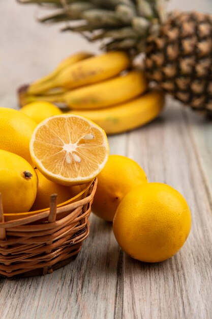 Zijaanzicht van rijk aan vitamines citroenen op een emmer met citroenen, bananen en ananas geïsoleerd op een grijze houten tafel op een witte muur