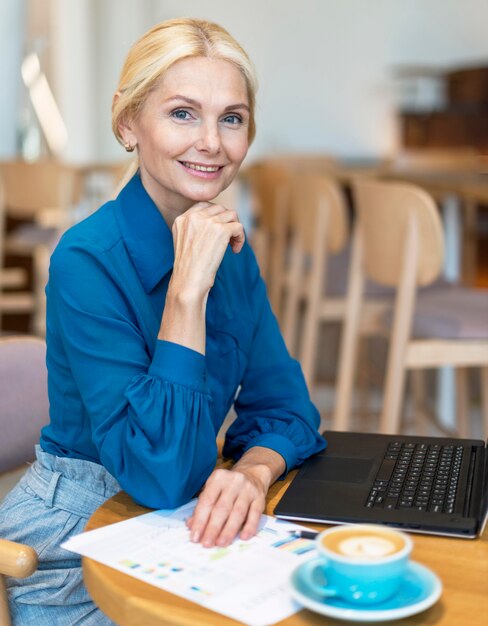 Zijaanzicht van smiley oudere zakenvrouw poseren tijdens het werken op laptop en koffie drinken