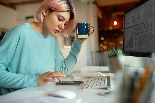 Zijaanzicht van stijlvolle roze harige jonge vrouwelijke blogger zittend aan tafel voor desktop computer