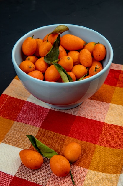 Gratis foto zijaanzicht van vers rijp kumquat fruit in een blauwe kom op geruite servet op zwarte ondergrond