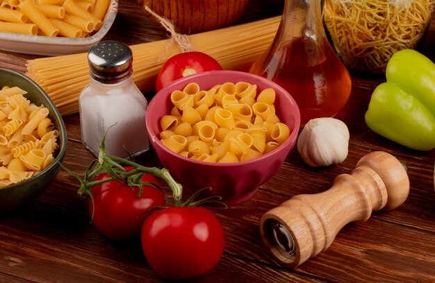 Zijaanzicht van verschillende macaronis en tomaten knoflook peper zout en gesmolten boter op houten tafel