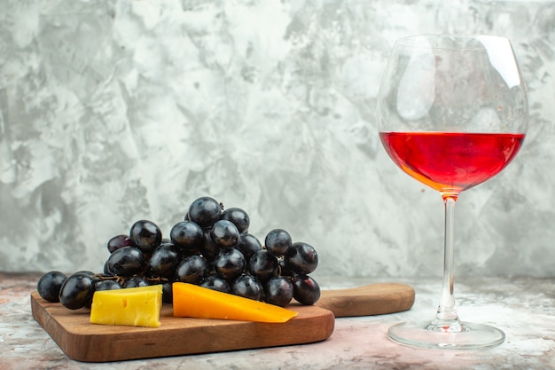 Zijaanzicht van verse heerlijke zwarte druiventros en kaas op houten snijplank en een glas wijn op gemengde kleurenachtergrond