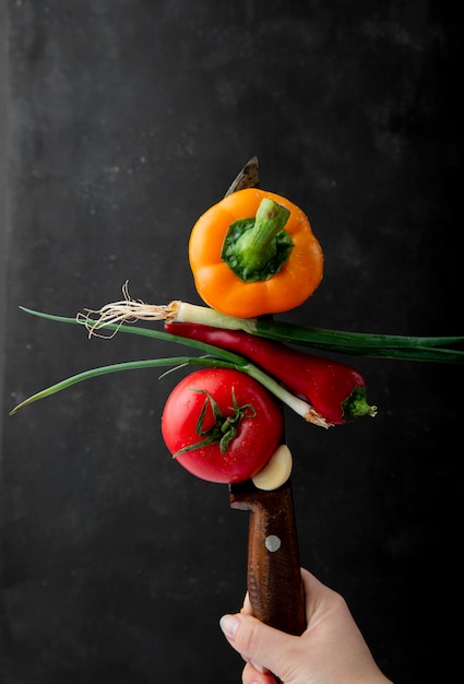 Zijaanzicht van vrouwelijke hand met een mes met verse rijpe groenten gele paprika groene ui rode chili peper en tomaat op zwarte achtergrond
