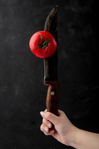 Zijaanzicht van vrouwelijke hand met rijpe natte tomaat met mes op zwarte achtergrond
